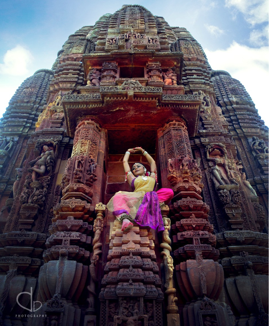 Kaustavi Sarkar poses in a brightly colored Odissi performance costume with arms raised over her head, 
   balanced in a niche of the Rajarani Temple in Bhubaneswar, Odisha, India. She is surrounded by sculptural figures likewise
   posed as dancers; the same image is both the focal point of the homepage and (in enlarged form) its background.