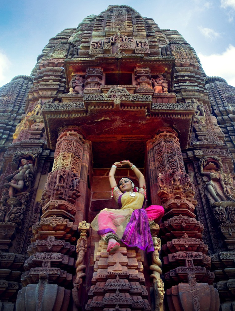 Kaustavi Sarkar is posed in a brightly colored Odissi performance costume with arms raised over her head, 
   balanced in a niche of the Rajarani Temple in Bhubaneswar, Odisha, India. She is surrounded by sculptural figures likewise
   posed as dancers; the same image is both the focal point of the homepage and (in enlarged form) its background.