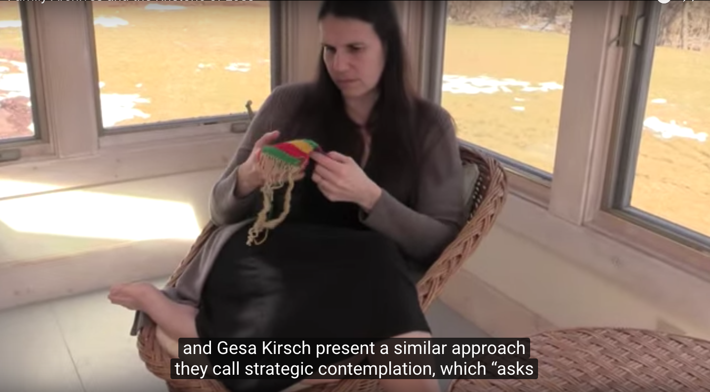 image of the author sitting cross-legged in a wicker chair in a sunlit room with windows behind her; she holds a small colorful 
piece of roughly woven yarn and looks at it intently